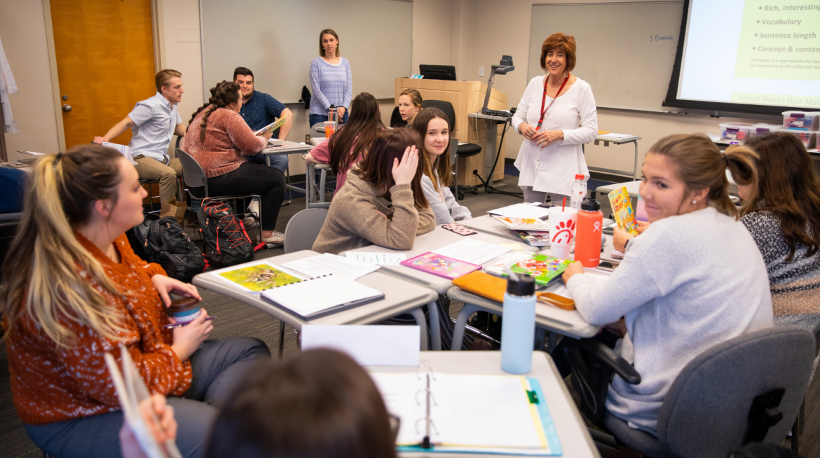 A CEHHS student just below the camera lens and out of focus holds up a children's book while classmates around them and Joan Grim look intently. Around the room, other student in groups talk amongst each other.