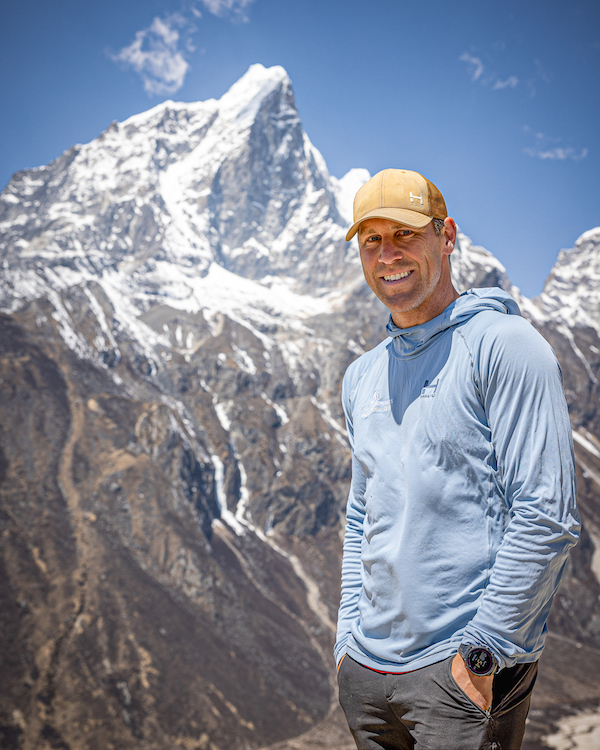 Bryan Hill stands with Mount Everest's peak in the distance. He has tan skin and is wearing a baby blue hooded sweatshirt and a baseball hat.