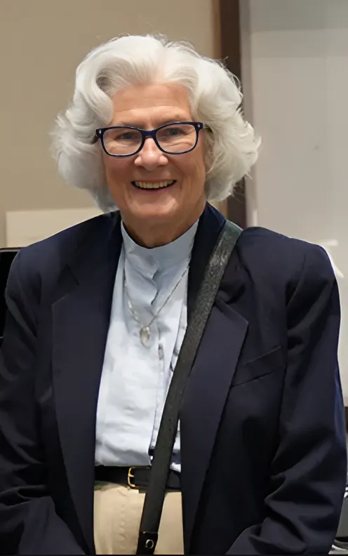 Portrait of Charmaine Mamantov—an older woman with light skin, black glasses, and chin-length curled white hair—wearing a pale blue button-down shirt and a navy blazer, while standing in a beige room.