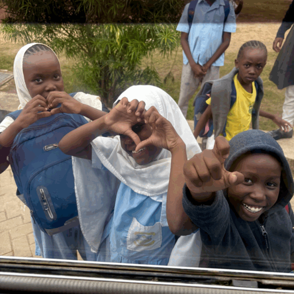 Kenyan children smile at the camera while standing outside a window, looking into an elementary school. The little girl in the middle is making a heart with her hands.