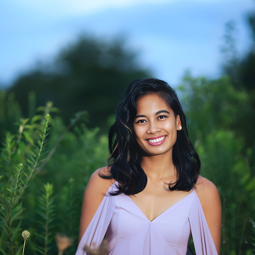 A portrait of Kirsten Salonga—a woman with deep tan skin and chest-length black hair—standing outside, wearing a lavender blouse with drapy sleeves that expose her shoulders.