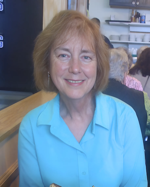 Portrait of Sarah DeYoung—an older woman with light skin and chin-length light brown hair—smiling while sitting in a restaurant, wearing a light blue button-down shirt, with a warm, professional expression.
