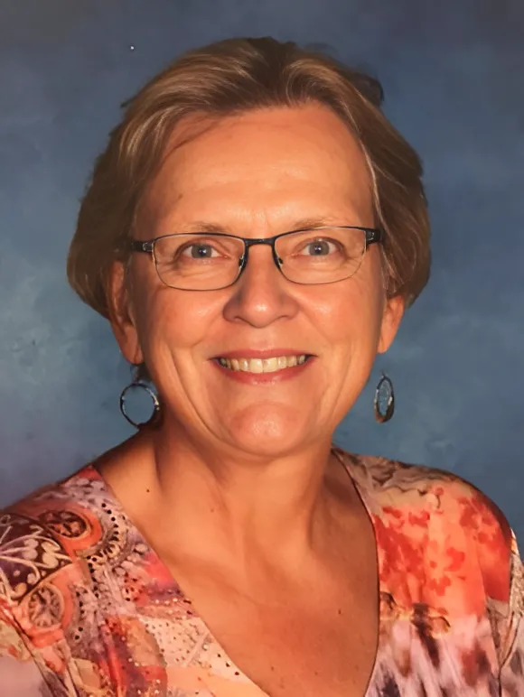 Portrait of Susan Espiritu—an older woman with tan skin, short dark blonde hair—wearing a floral blouse and smiling gently toward the camera against a blue background.