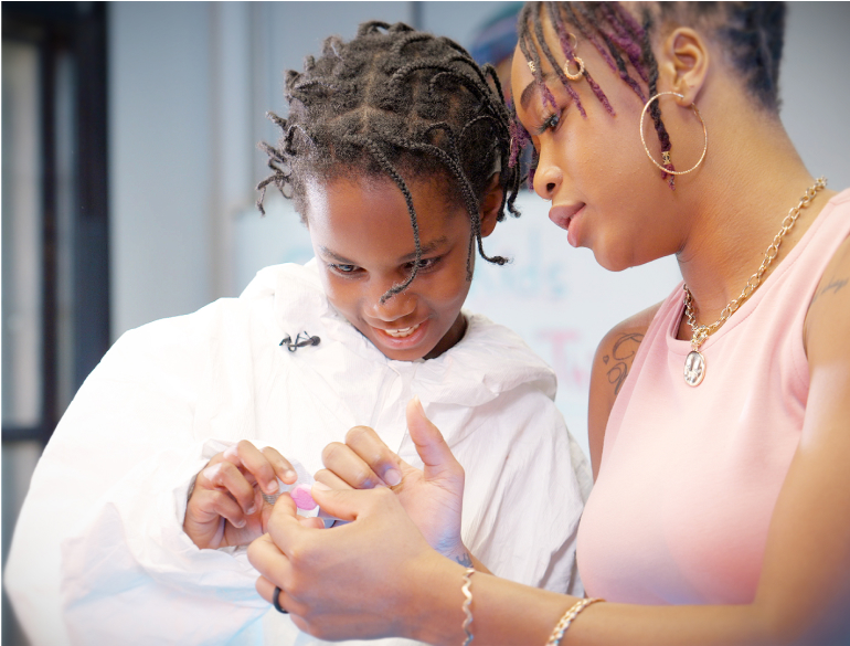 A young child with dark brown skin and short, twisted black hair smiles while carefully examining a small electronic component. An adult with medium-brown skin leans in beside the child, offering guidance. The adult has dark hair with burgundy highlights styled in twists pulled into an updo and wears large gold hoop earrings, layered gold necklaces, and a sleeveless light pink top. Both focus intently on the hands-on activity in a classroom setting.